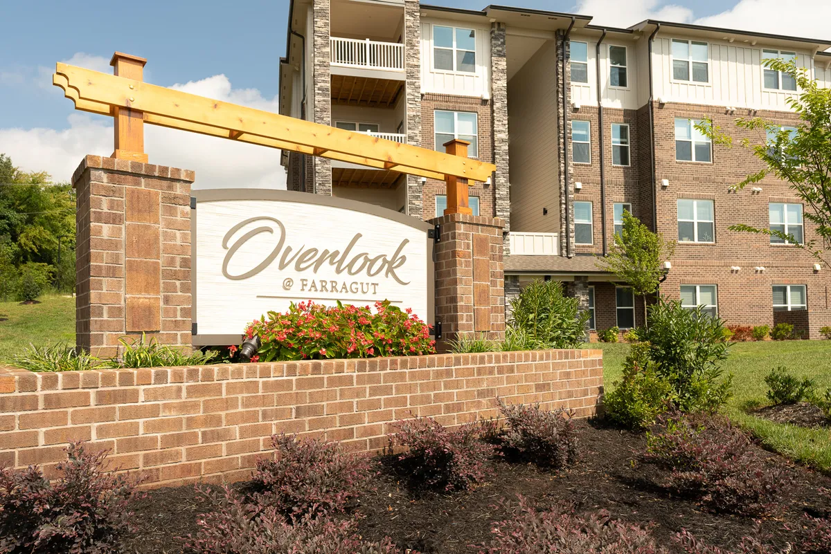 The image shows inviting signage, lush landscaping, and the attractive building facade. Residents are seen walking and relaxing, highlighting the well-maintained communal spaces and an inviting, community-focused atmosphere.