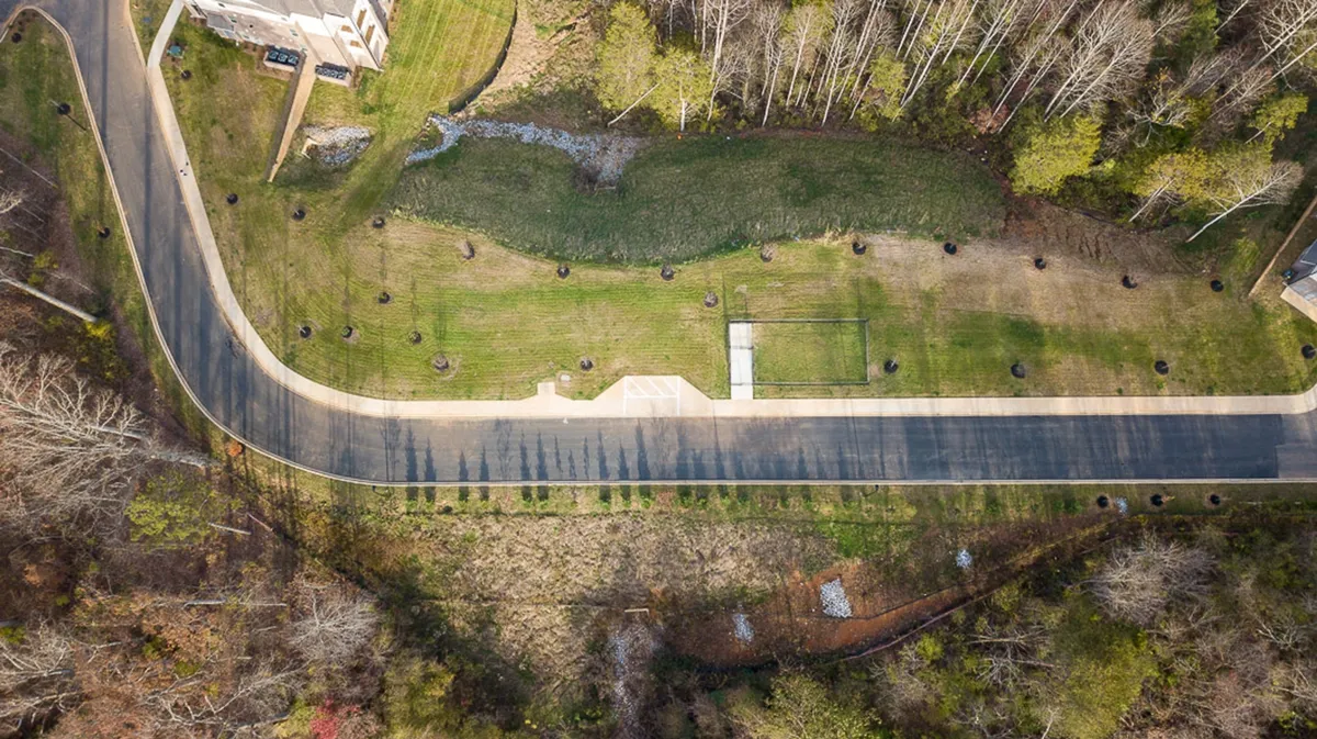 A gently curving paved road is bordered by a neatly manicured lawn and mature trees, creating an inviting environment for walking or jogging. The peaceful mood and visible greenery reinforce accessibility to nature and outdoor spaces.
