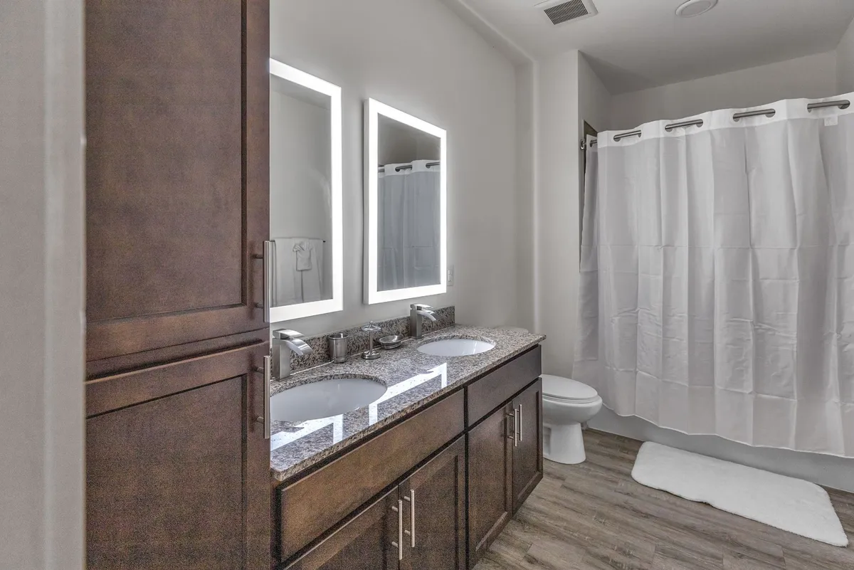 A modern bathroom with double sinks, large mirrors, a sleek granite countertop, and a subtle shower curtain in the background. The fixtures and finishes are contemporary and spacious, evoking a sense of cleanliness and comfort.