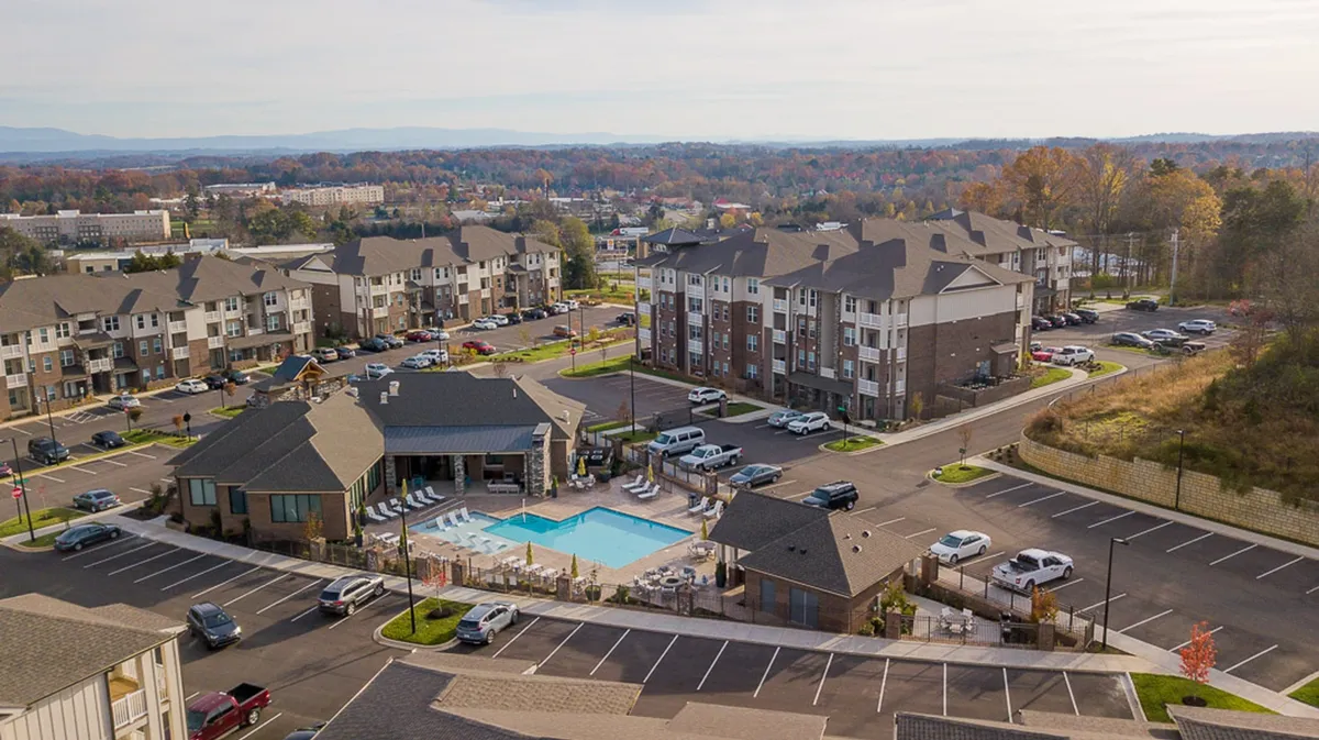 This exterior scene highlights the resort-style swimming pool in the foreground, flanked by modern residential buildings and a visible parking area. The setting is active with residents swimming, sunbathing, and socializing poolside, all beneath a warm sunset with mountain views in the background.