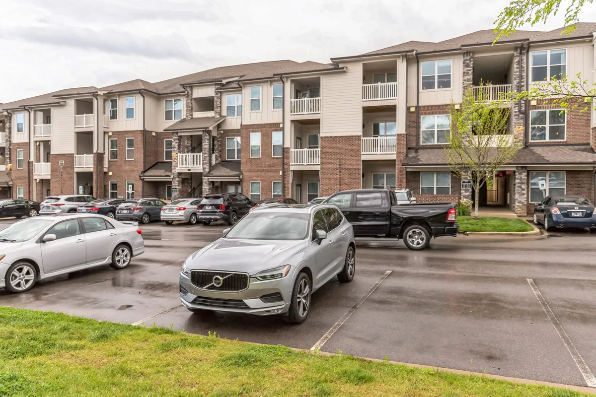 This exterior scene presents modern apartment buildings framed by green lawns and parked vehicles. Residents are seen mingling casually in the parking lot, suggesting an active, welcoming community. The open green space is pet-appropriate, supporting outdoor activities like morning walks.