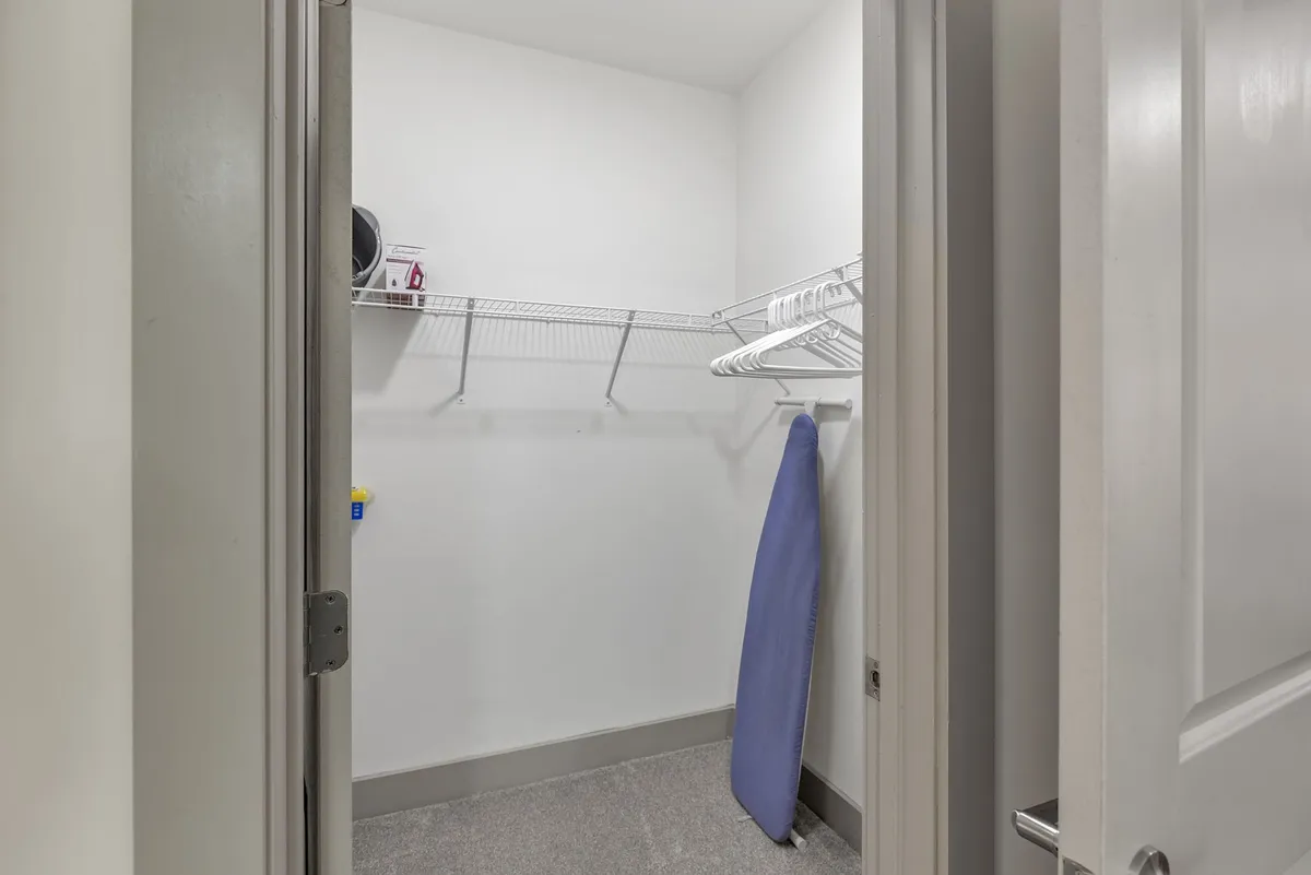 A practical laundry and storage area inside the apartment, featuring wire shelving and a blue ironing board. The space is organized, emphasizing easy access and efficient use of in-unit amenities.