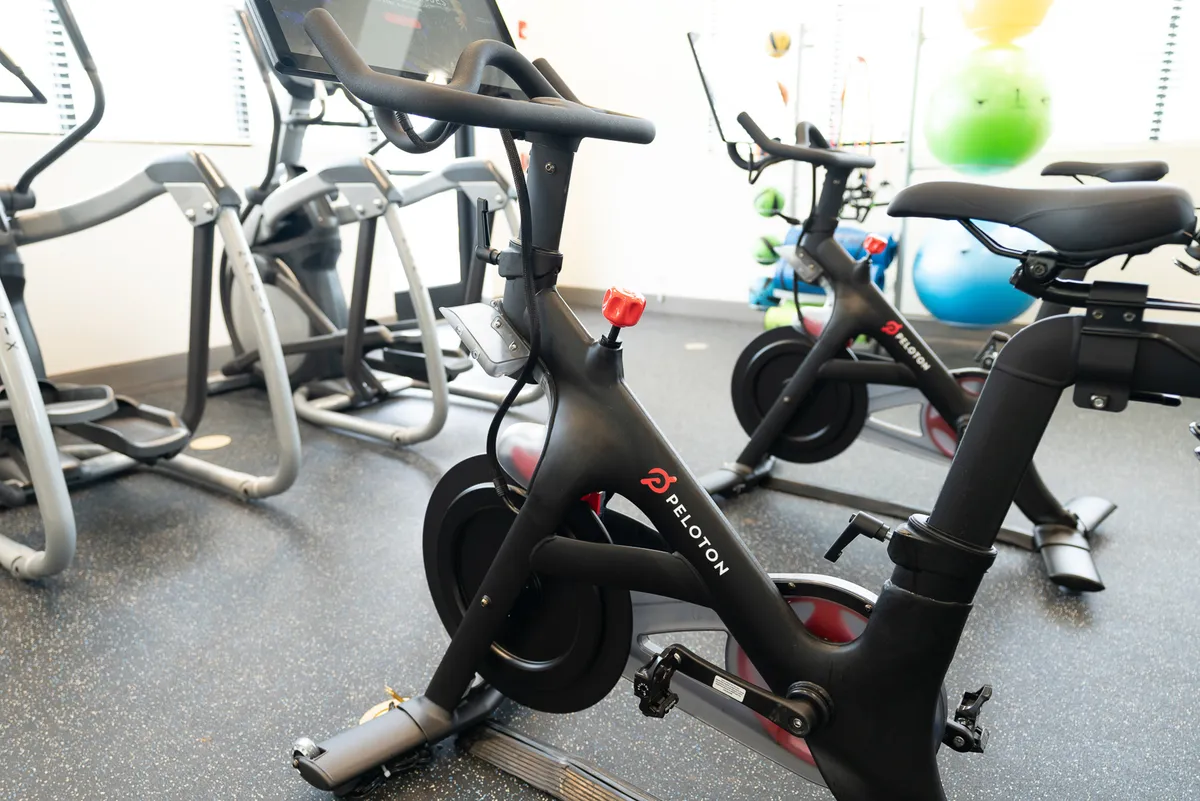 A bright, modern fitness center interior featuring a Peloton bike, elliptical machines, and colorful exercise balls. The space is designed for motivating workouts, with plenty of natural light and energetic vibes.