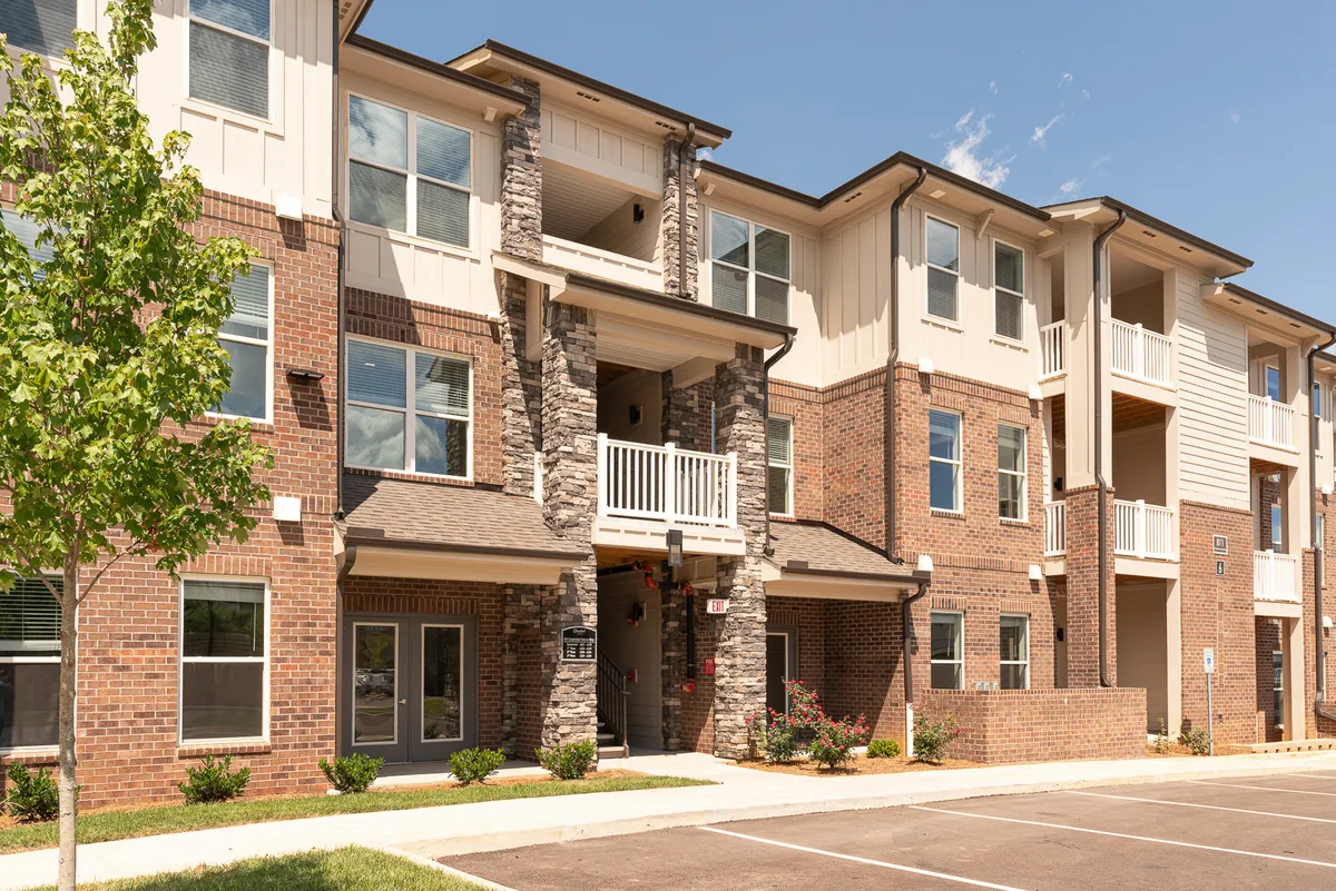This exterior shot captures the building's stone facade, large windows, balconies, and well-maintained green landscaping. People can be imagined enjoying coffee on balconies or relaxing in the communal yard, reflecting everyday comfort and connection to the outdoors.