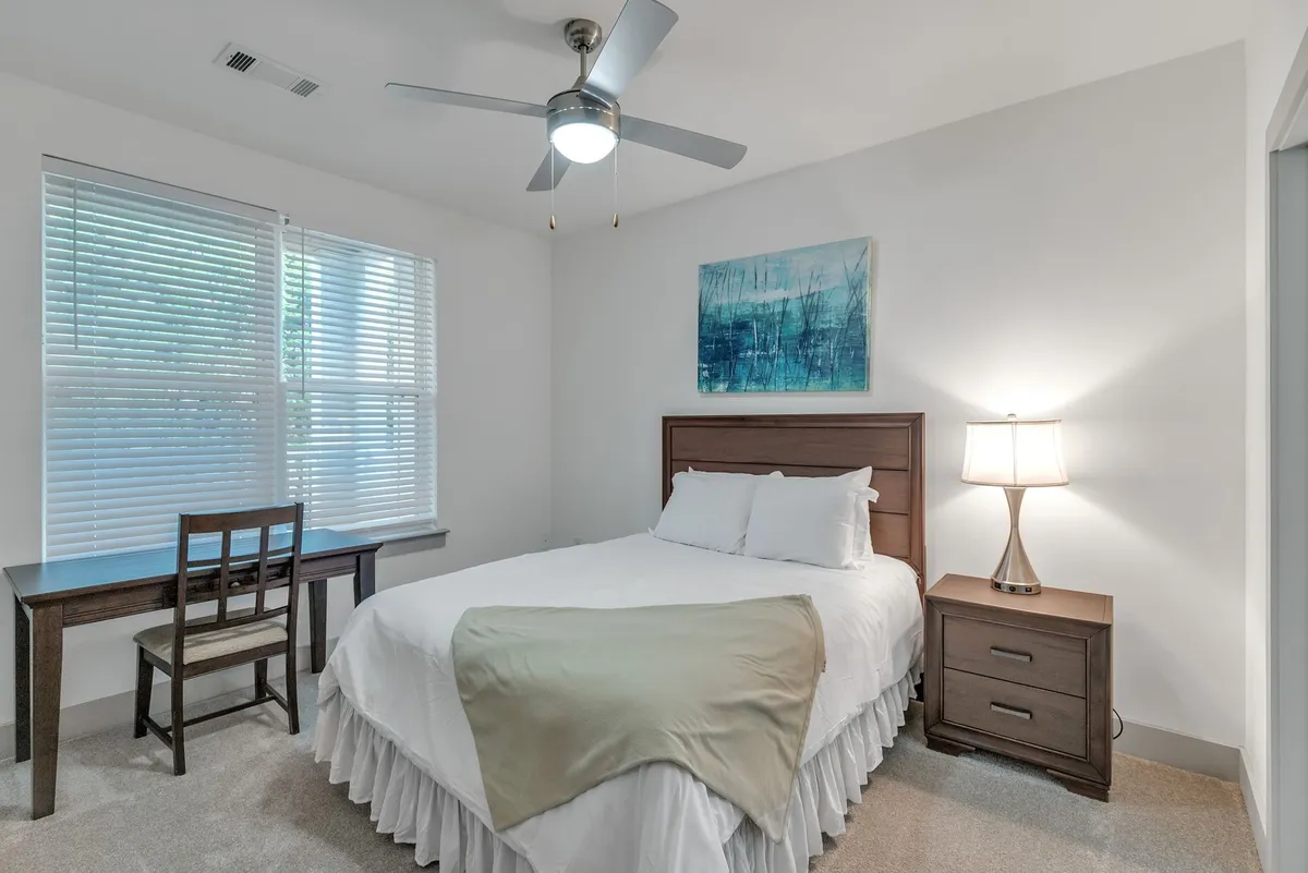 The image showcases a tranquil, sunlit bedroom with a plush queen bed, a modern desk setup for work or creative pursuits, soft ambient lighting from a stylish lamp, and elegant artwork adorning the walls. The overall mood is serene and inviting, signaling both comfort and productivity.