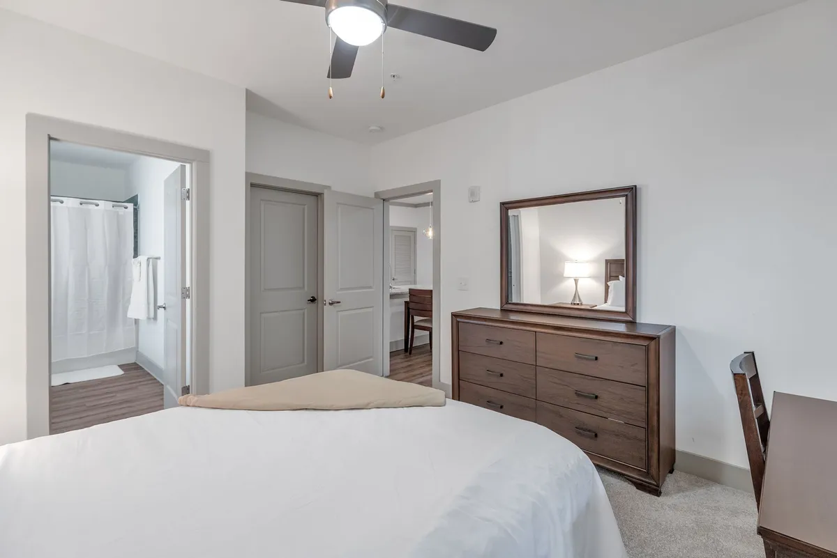 A tranquil bedroom featuring a bed made up in crisp white linens, a wooden dresser with a mirror, a small desk and chair, and an open door offering a glimpse into the adjacent bathroom. The space is softly lit, creating a restful ambiance ideal for relaxation or preparing for the day.