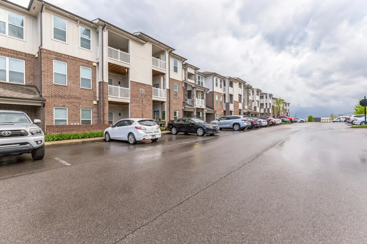 This image features the apartment complex's exterior with visible walkways, parked cars, and landscaped areas. The mood is inviting and peaceful, and the walkways suggest a great place for daily strolls with pets. The surrounding green spaces also hint at proximity to greenways for outdoor activities.