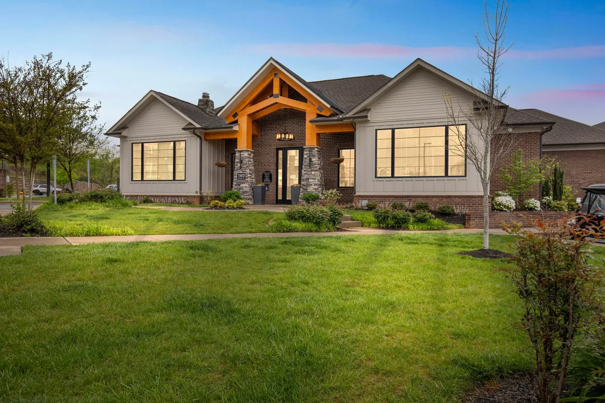 The exterior of the building features a bold orange entryway, large windows, stone accents, and a manicured lawn. The scene captures social gatherings, family barbecues, and evening strolls, giving a sense of community and connection to local life.