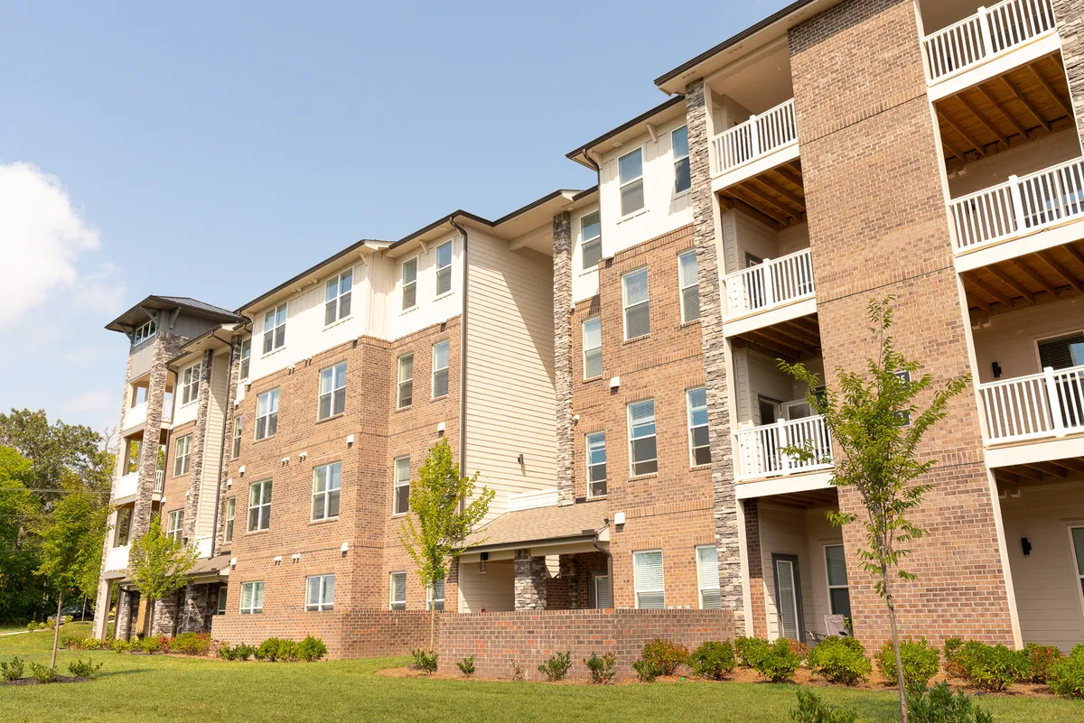A wide-angle view of modern brick buildings with balconies overlooking landscaped gardens. The scene captures the vibrancy of outdoor living, with people relaxing on balconies and walking in the landscaped yard, evoking a pet- and family-friendly atmosphere.
