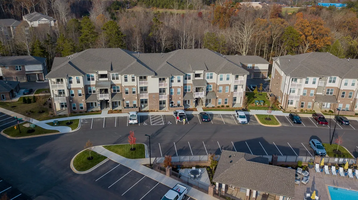 A wide-angle exterior shot featuring apartment buildings surrounding a landscaped swimming pool and a spacious parking lot. Residents are seen enjoying outdoor leisure and gathering in common spaces, with well-kept greenery enhancing the inviting ambiance.